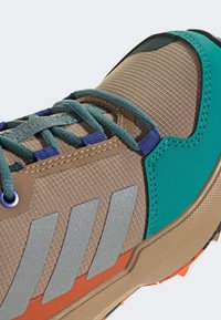 Beige textilsneaker med teal- och orange-accenter, med reflekterande silvriga ränder, intrikat textur och blå snören.