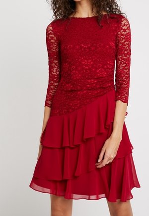 Femme portant une robe rouge mi-longue avec un corsage en dentelle, des manches trois-quarts, et une jupe à volants superposés, se tenant devant un fond uni.