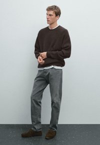 Bruine geribbelde trui, grijze jeans en bruine suède schoenen. Het model staat tegen een lichtgrijze achtergrond.
