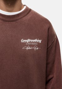 Felpa marrone con scollo a girocollo a coste, presenta il logo bianco "Goodfornothing" e la firma sul petto, con una texture di cotone morbido.