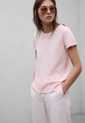 Młoda kobieta w jasnoróżowej koszulce i białych spodniach, z rękami w kieszeniach, pozująca na jednolitym szarym tle.