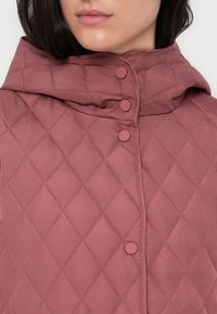 Αμάνικο καπιτονέ μπουφάν σε ροζ σκόνη ύφασμα με ψηλό γιακά. Διακριτικά χαρακτηριστικά περιλαμβάνουν στρογγυλεμένα κουμπώματα και λεία υφή.