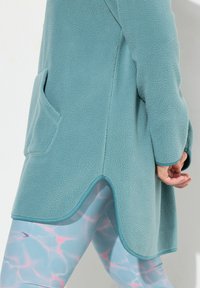 Valkoinen teal fleece-takki, jossa on teksturoitu pinta ja sivutaskut, yhdistettynä vaaleansinisiin legginsseihin, joissa on vettä inspiroiva vaaleanpunainen kuvio.