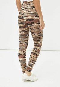 Persona vistiendo leggings de estampado abstracto en marrón, beige y negro, y zapatillas blancas, de pie con una mano relajada a su lado.