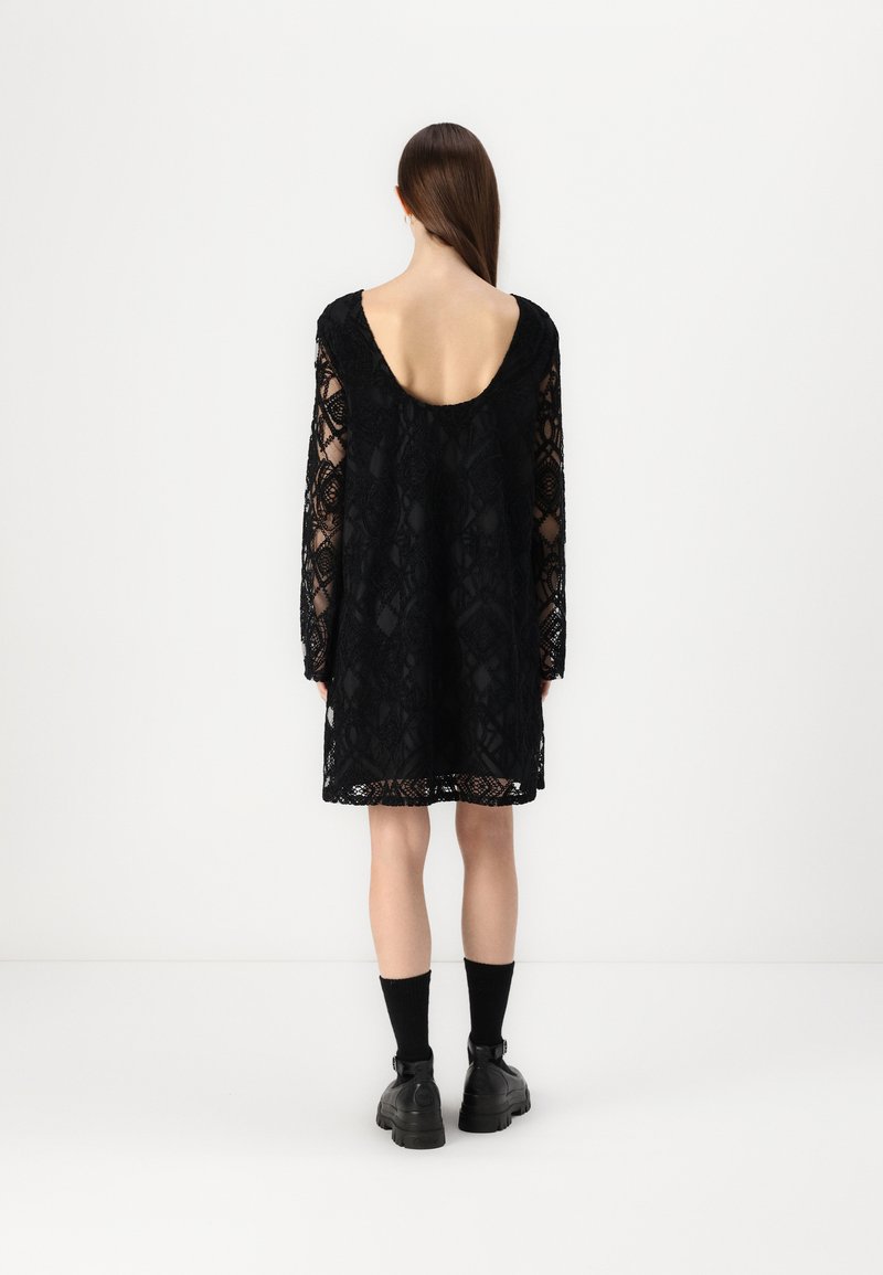 Vestido negro de encaje con mangas largas, escote en la espalda y patrones geométricos intrincados. Combinado con botas negras hasta el tobillo y calcetines.