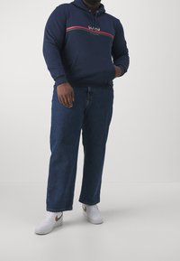 Sudadera marinera con un bolsillo frontal, detalles en franjas blancas y verdes, combinada con jeans rectos azul oscuro y zapatillas blancas con logo dorado.