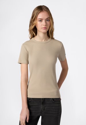 Giovane donna con capelli biondi sciolti che indossa una maglietta a maniche corte beige a coste e jeans neri, in piedi contro uno sfondo bianco.