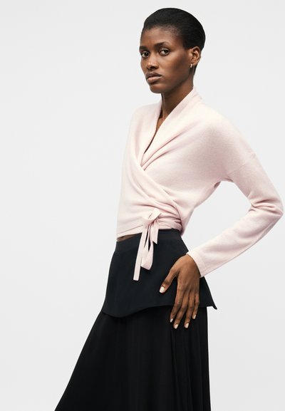 Kobieta o ciemnej karnacji, ubrana w jasnoróżowy sweter z wiązaniem w pasie oraz czarną warstwową spódnicę, stojąca na jednolitym tle.