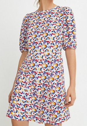 Kobieta nosząca białą sukienkę z krótkim rękawem z kolorowym kwiatowo-owocowym wzorem, bufkami i okrągłym dekoltem.