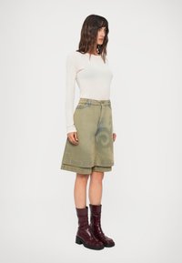 Světle dlouhý rukávový top, doplněný zelenými džínovými šortkami ke kolenům s vírovým designem. Modelka má na sobě lesklé bordó kotníkové boty.