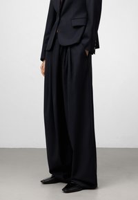 Persona che indossa pantaloni neri su misura dal taglio ampio, con un blazer nero abbinato e scarpe nere a punta quadrata, in piedi contro uno sfondo semplice.