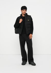 Black puffer vest with high collar and gold logo, worn over a black sweater and paired with loose black pants and textured black shoes.