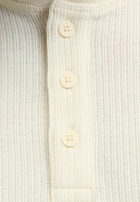 Kremfarget ribbestrikket stoff med en teksturert overflate, med fire knappelukkinger og runde knapper med fire hull hver.