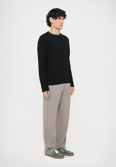 Czarny sweter z dzianiny z okrągłym dekoltem, szare plisowane spodnie i zielono-szare sneakersy z białymi sznurówkami, noszone przez męskiego modela.