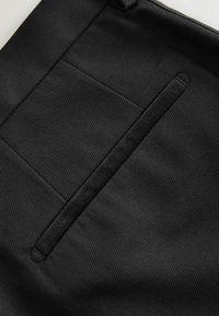 Schwarzer Stoff mit einer strukturierten, gerippten Oberfläche. Enthält eine horizontale Tasche mit einem schmalen, dunklen Akzent. Sauberes, minimalistisches Design.