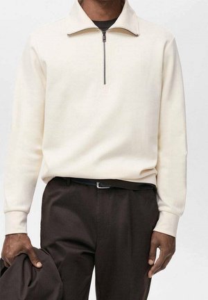 Mann trägt cremefarbenen Pullover mit Viertel-Reißverschluss, in dunkelbraune Hose mit schwarzem Gürtel gesteckt, hält dunkelbraunen Stoff in der linken Hand.
