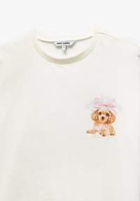 Cremefarvet T-shirt med en blød tekstur, som har et trykt billede af en hvalp iført en lyserød sløjfe, ledsaget af tekst i farverig skrifttype.