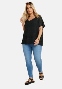 Túnica negra de punto abierto con mangas cortas; combinada con jeans ajustados azules y sandalias planas negras con un detalle de hebilla.
