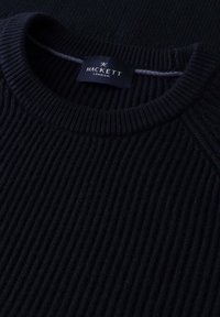 Gros plan d'un pull noir à col rond en maille côtelée avec une étiquette Hackett London bleu foncé cousue à l'intérieur du col.