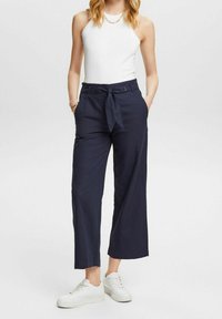 Navy vida byxor med knytmidja, med sidofickor och en slät textur, ihop med en vit ärmlös topp och vita sneakers.