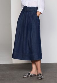 Denim midi-rok in donkerblauw met een hoge taille, wijde plooien en een bijpassende riem. Gecombineerd met zwart-witte slip-on schoenen met patroon.