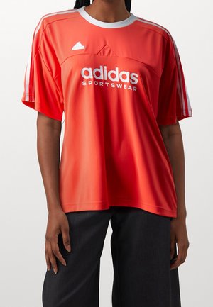 Person iført en løs rød Adidas sportstrøje med hvide striber på ærmerne og sorte bukser, stående foran en ensfarvet baggrund.