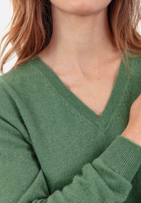 Pull vert en maille avec col en V, poignets côtelés et texture douce. Gros plan montrant le détail du tissu et du design de l'encolure.