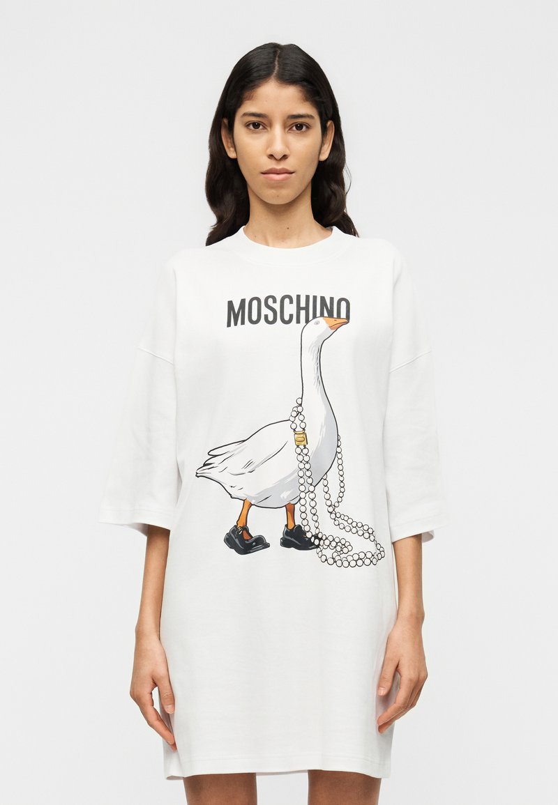 Λευκό, υπερμεγέθες φόρεμα T-shirt με γραφικό εικόνας μιας χήνας με μαύρα παπούτσια και κολιέ από μαργαριτάρια. Εμφανίζεται λογότυπο στο πάνω μέρος.