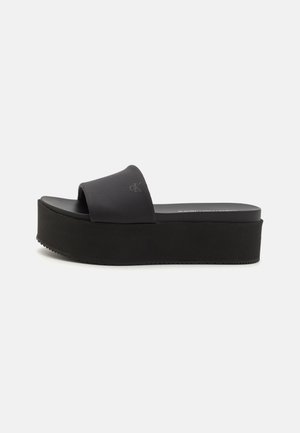 Black platform slide sandal with a thick black rubber sole, textured upper strap, and minimalistic design, featuring a subtle brand logo.
