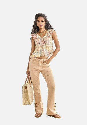 Mujer con top sin mangas floral con volantes, pantalones beige con lazadas laterales, sandalias y sosteniendo un bolso tejido con asas de cuero.