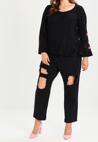 Haut noir à manches longues avec broderie florale sur les manches, associé à un jean noir déchiré avec de grandes déchirures, et des talons hauts roses.