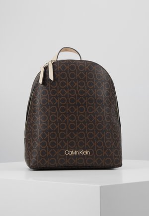Brązowy plecak Calvin Klein z powtarzającym się wzorem "ck" i złotym logo, beżowym uchwytem oraz podwójnymi zamkami błyskawicznymi na białej powierzchni.
