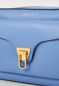 Borsa a tracolla in pelle blu chiaro con superficie testurizzata, chiusura in metallo dorato e bordi neri cuciti. Logo impresso sul davanti.