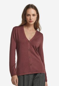 Kvinna med ljusbrunt hår som bär en knappad, långärmad, V-ringad vinröd cardigan och grå texturerade byxor mot en vit bakgrund.