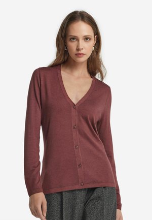 Kvinna med ljusbrunt hår som bär en knappad, långärmad, V-ringad vinröd cardigan och grå texturerade byxor mot en vit bakgrund.