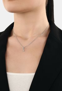 Silverkedja med en liten cirkulär hängsmycke med en klar ädelstensaccent, bärs över en vit topp och under en svart blazer.