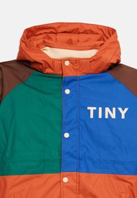 Veste colorée pour enfants en orange, bleu, vert et marron avec une capuche et des boutons-pression blancs, affichant le mot "TINY" sur la poitrine.