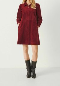 Robe en velours côtelé bourgogne avec de longues manches bouffantes, fermeture éclair centrale et poches latérales. Des bottes noires jusqu'aux genoux complètent la tenue.