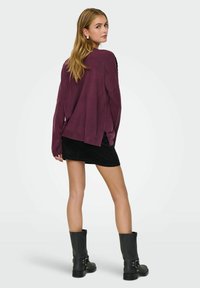 Übergroßer burgunderroter Pullover mit langen Ärmeln; getragen über einem schwarzen Cord-Minirock und schwarzen kniehohen Stiefeln. Texturiert, mit seitlichen Schlitzen.