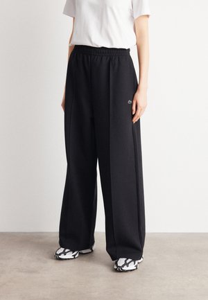Personne portant un pantalon noir à jambes larges avec une taille élastique et des baskets blanches, debout sur un sol gris contre un mur blanc.