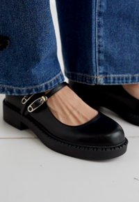 Black leather Mary Jane shoes with a round toe, flat sole, and a chunky design. Features silver buckle hardware and raised stitching details.