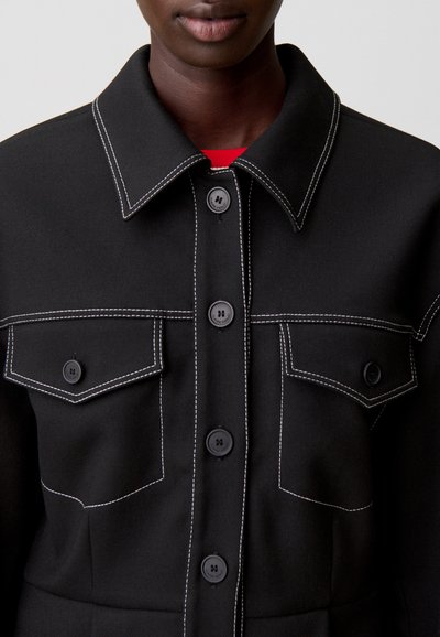 Giacca nera con collo a risvolto, caratterizzata da cuciture bianche e due tasche al petto. Chiusura con bottoni neri sulla parte anteriore.