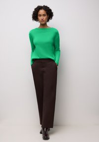 Pull vert en tricot avec un large décolleté, associé à un pantalon large marron et des bottes sombres. Le mannequin pose les mains dans les poches.