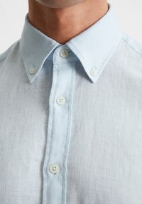 Camicia in lino azzurro chiaro, con colletto e tre bottoni frontali allacciati, indossata da una persona.