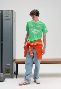 Grünes Sporttrikot mit weißem Logo, lässig sitzende blaue Jeans, orangefarbener Pullover in der Taille gebunden und beige Sneakers mit roten Streifen.