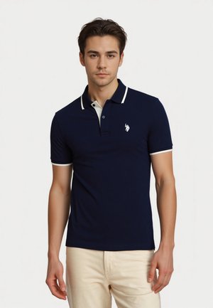 Young man wearing a navy polo shirt with light collar and embroidered logo, standing against a plain white background.