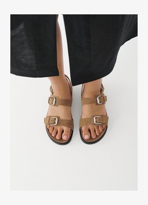 Pieds portant des sandales en daim brun avec des boucles argentées, posés sur une surface blanche, avec un pantalon noir visible au-dessus des chevilles.