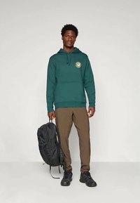 Moški v zeleni kapuci z okrožnim logotipom, rjavih hlačah in črnih čevljih, ki drži črn nahrbtnik z eno roko, stoji ob preprosti ozadju.