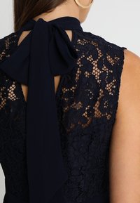 Nära bild av en ärmlös klänning i marinblå spets med blommönster och en stor knytfluga vid halsen, bärs av en person med stora hoopörhängen.