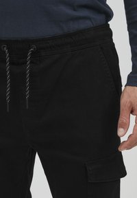 Černé cargo kalhoty s elastickým páskem na stahování, s detailním prošíváním a boční kapsou. Materiál vypadá hladce s mírným leskem.
